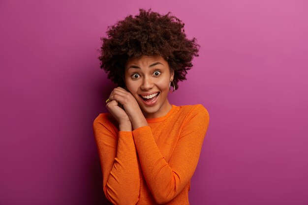 Isolated shot of pretty ethnic young woman keeps hands near face, smiles pleasantly, being in good mood, dressed in casual orange jumper, has natural curly hair, poses against purple wall