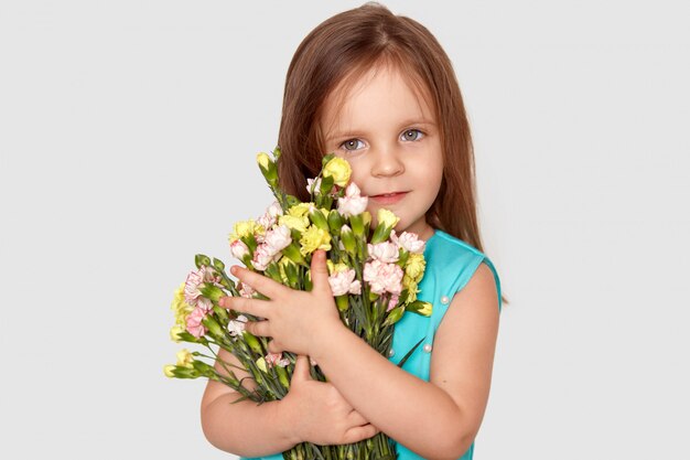Изолированный снимок довольно маленькой девочки готовит букет цветов к маме на День Матери, имеет привлекательный внешний вид, одет в праздничную одежду, изолирован на белой стене. Концепция весны и детей