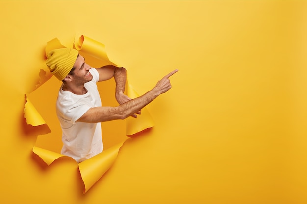 Isolated shot of satisfied male model stands sideways in paper hole, dressed in yellow headgear