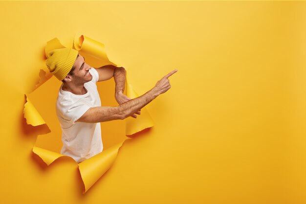 Isolated shot of satisfied male model stands sideways in paper hole, dressed in yellow headgear