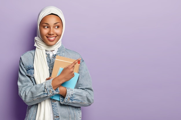 Isolated shot of satisfied Muslim student shows awesome advert, points at upper right corner, wears white veil and jean jacket