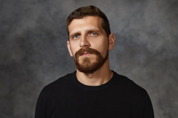 Isolated shot of serious handsome thirty year old Caucasian man with blue eyes, mustache and thick beard posing against blank wall ,, dressed in black sweater