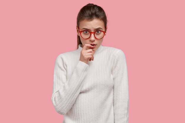 Изолированный снимок серьезной приятной на вид женщины держит указательный палец возле губ, серьезно смотрит через очки, одетая в белую повседневную одежду