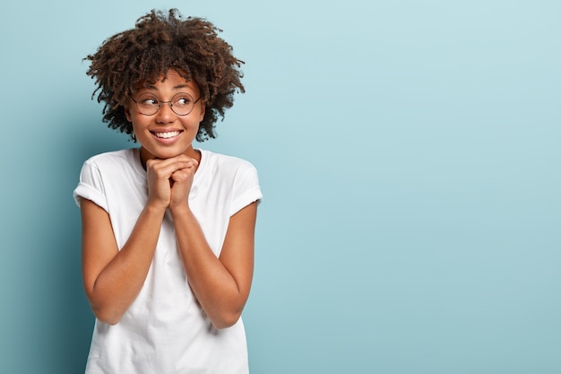 Isolated shot of smiling dark skinned teenager keeps hands under chin, looks aside