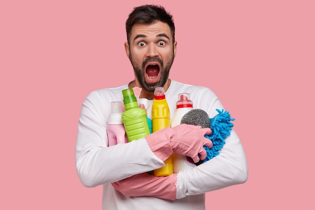 Isolated shot of stupefied bearded emotional man carries bottles of detergents closely, stares with unbeliveable expression, dressed casually