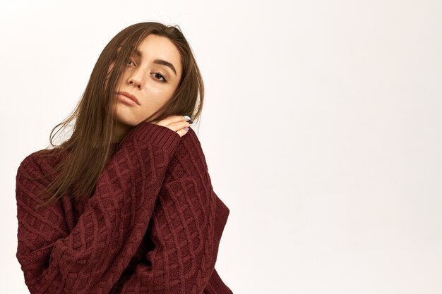 Isolated shot of stylish cute young woman freezing from cold because heating turned off, trying to warm up in knitted sweater, hugging herself