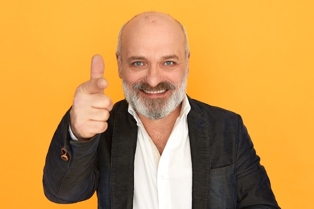 Isolated shot of successful positive friendly senior businessman in formal elegant clothes smiling pointing fore finger at camera, recruiting you, offering position in his company. body language