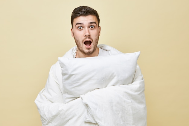 Isolated shot of terrified young bearded male rolling himself in white blanket and shouting with mouth wide opened, can't fall asleep because of scary movie. Bedtime, sleep and insomnia concept