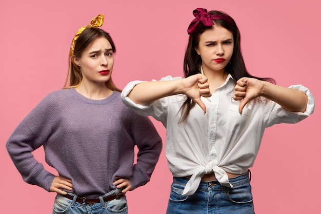 Free photo isolated shot of two attractive young woman expressing disapproval, dislike, being disappointed with bad quality, showing thumbs down gesture, having displeased looks.