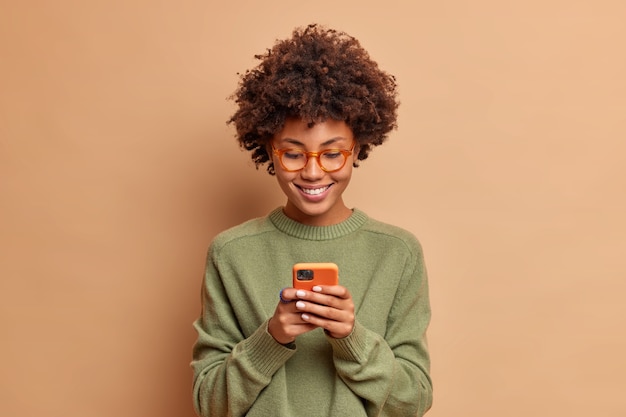 Isolated shot of woman uses smartphone application enjoys browsing social media creats news content makes online order wears spectacles and casual jumper poses over beige studio wall