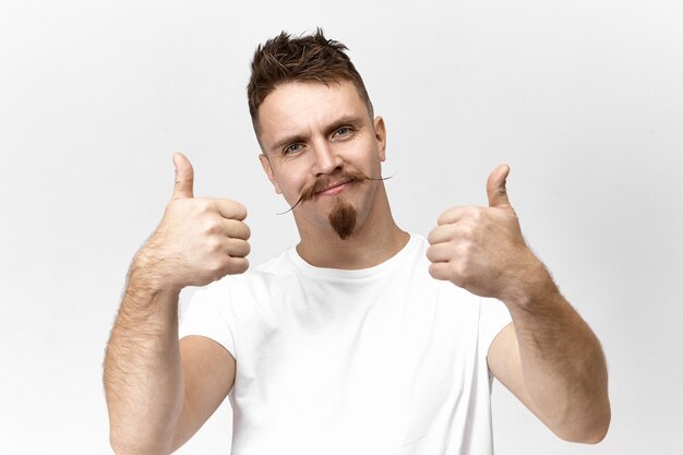 Isolated studio shot of handsome trendy young Caucasian man with goatee beard and handlebar mustache looking at camera with positive friendly smile, showing thumbs up sign, liking idea or plan