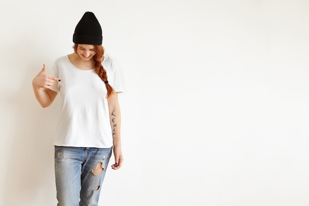 Isolated studio shot of young female with braid looking down as she pointing at her blank white t-shirt