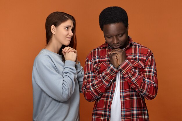 Free photo isolated unhappy depressed young interracial couple white woman and black man facing financial or health problems, praying, holding hands clasped, having mournful facial expressions