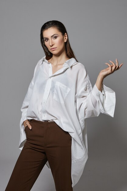 Isolated vertical shot of stylish young brunette female in trendy clothes having serious confident facial expression, gesturing as if holding something in open hand. Style, fashion and beauty