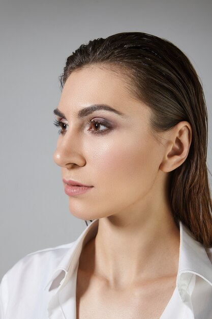 Isolated vertical view of fashionable stylish young brunette woman with male up and dark hair combed back posing, dressed in silk white shirt. Beauty, style, skin and hair care concept