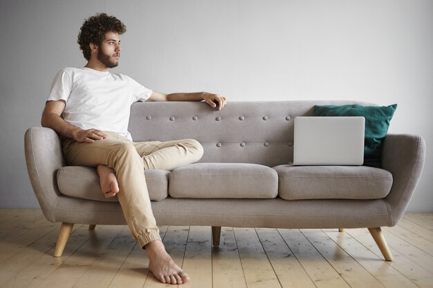 Isolated view of thoughtful young man with bare feet sitting indoors on sofa with open generic laptop, having small break while working distantly from home. People, job, occupation and technology