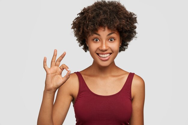 It is deal. Glad good looking woman with Afro hairstyle, dark skin, pleasant smile, shows okay or perfect gesture