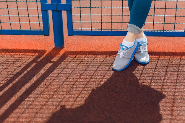 Это изображение ног девушки, стоящих у синего забора на стадионе. Она носит серые кроссовки с синей линией и синие брюки.