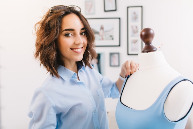 È un ritratto di una ragazza nello studio del laboratorio. ragazza castana con una camicia blu sta creando un vestito blu sul manichino. sta sorridendo alla telecamera.