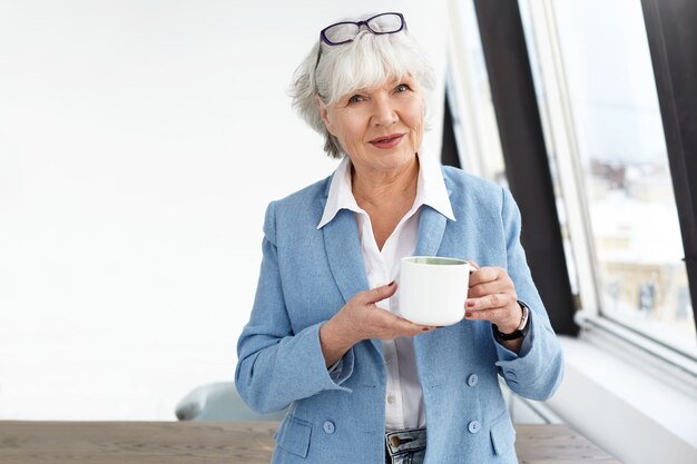 It's time for coffee break. Indoor image of elegant middle aged businesswoman wearing fashionable clothes and glasses holding white cup while drinking tea at her office, standing by window and smiling
