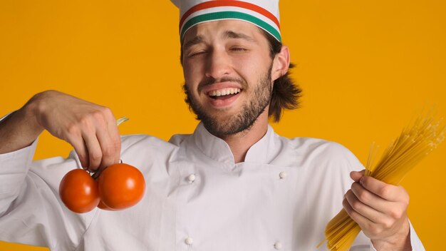 Italian chef dressed in uniform holding tomatoes and pasta in hands and singing over yellow background Emotional man in chef hat fooling around at work