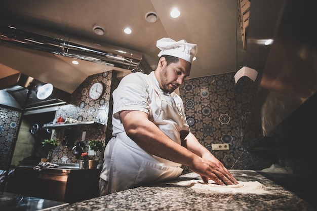 Italian chef in uniform is preparing pastry for pizza at the kitchen.