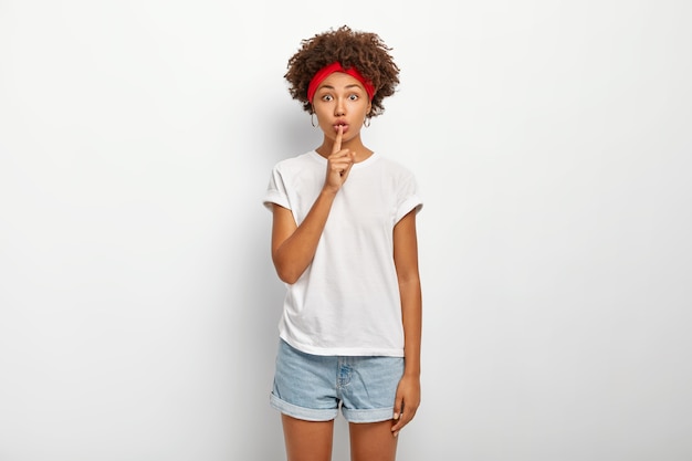 Free photo its better to keep quiet. intensed bothered woman has afro hairstyle, explains taboo, makes silence gesture, shushing sign, wears casual outfit, poses against white wall. keep your voice down