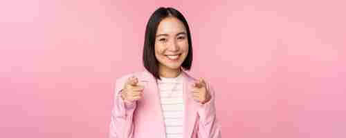 무료 사진 축하합니다 웃고 있는 열정적인 한국 기업 여성 사업가가 카메라를 가리키며 분홍색 배경 위에 서서 당신을 찬양하는 것을 축하합니다