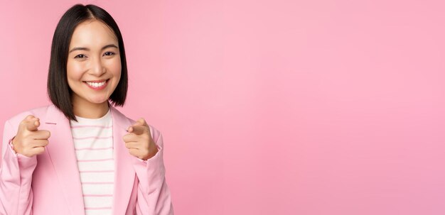 Это вы поздравляете Улыбающуюся восторженную корейскую корпоративную леди-бизнесвумен, указывающую пальцем на камеру, поздравляющую, хвалящую вас, стоящую на розовом фоне