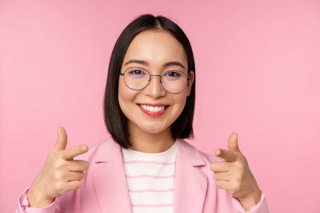 Its you congratulations Smiling asian corporate woman ceo manager in suit and glasses pointing fingers at camera recruiting praise or compliment standing over pink background