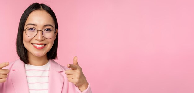 Это вас поздравляю Улыбающаяся азиатская корпоративная женщина-генеральный директор в костюме и очках, указывающая пальцем на камеру, вербующая похвалу или комплимент, стоя на розовом фоне