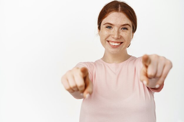 Its you. Smiling redhead girl pointing fingers looking happy, standing on white in pink t-shirt.