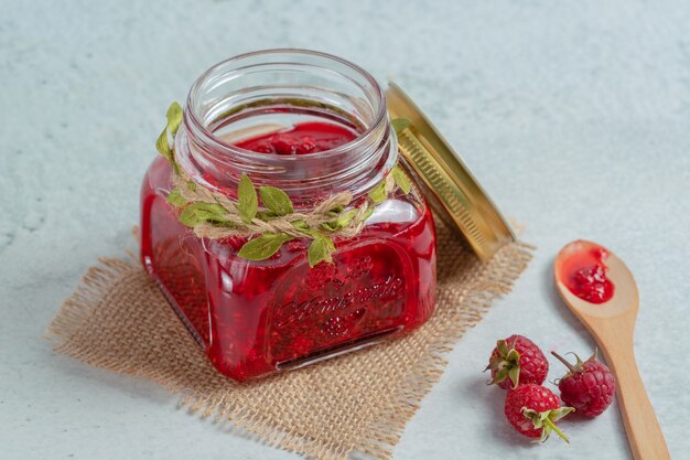 Jam in jar and raspberry on ground over grey surface.