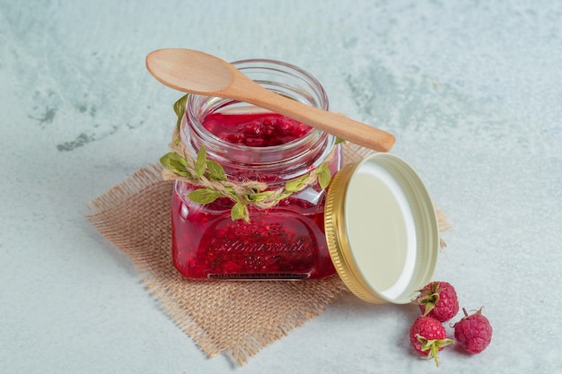 Jam in jar and raspberry on ground over grey surface. 