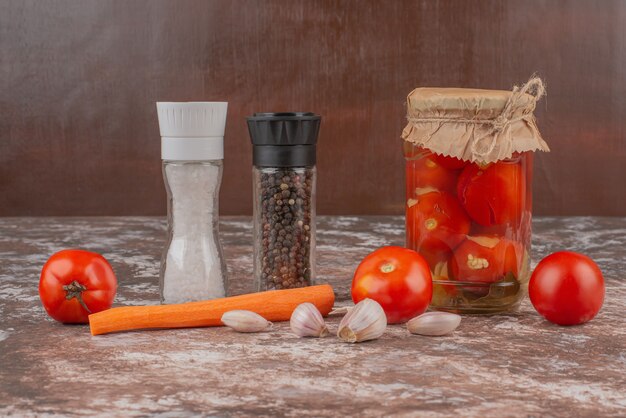 A jar of pickled tomatoes, pepper grains and fresh vegetables on marble table.