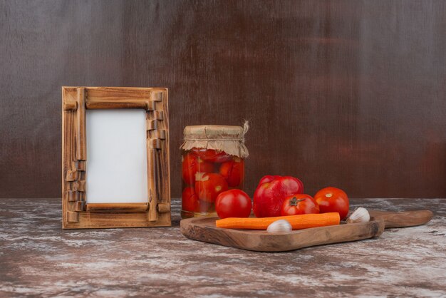 Jar of pickled tomatoes, plate of fresh vegetables and picture frame on marble table.