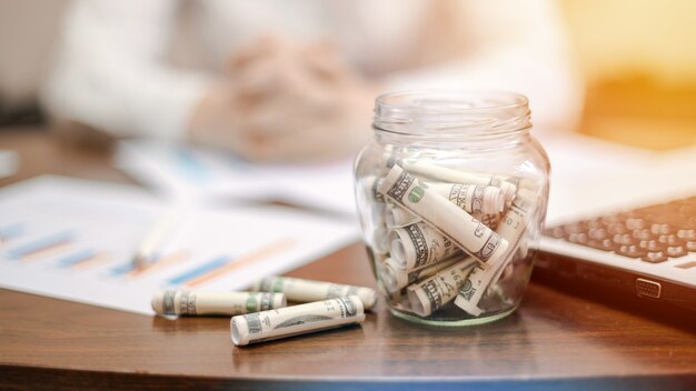 A jar with rolled banknotes on the table. Laptop, papers, woman on the background