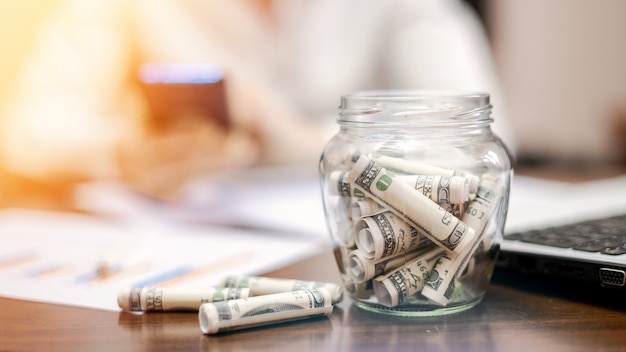 A jar with rolled banknotes on the table. Laptop, papers, woman on the background