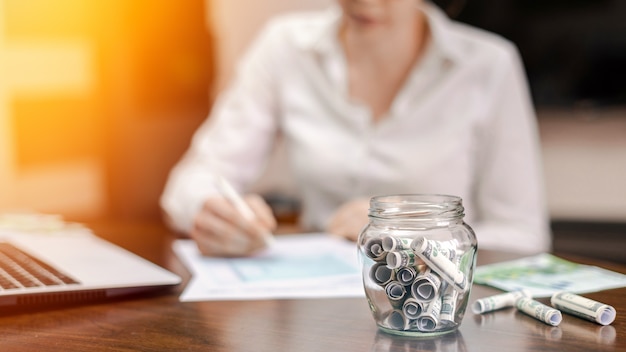 A jar with rolled banknotes on the table. Laptop, papers, woman on the background