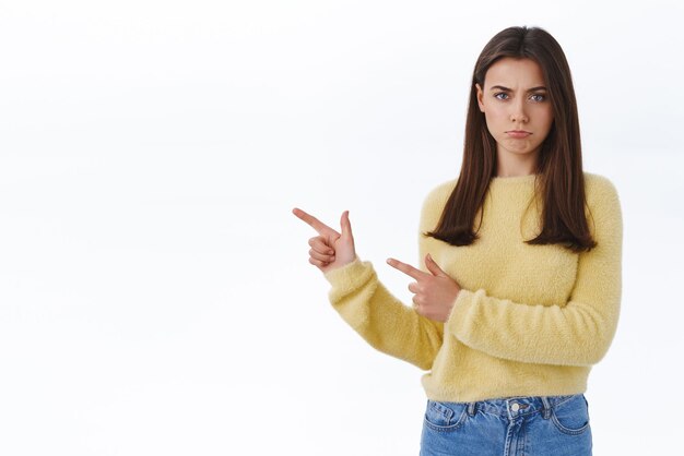 Jealous pouting offended cute sulky girlfriend in yellow sweater frowning and looking insulted as pointing finger left didnt receive something she wanted express disappointment white background