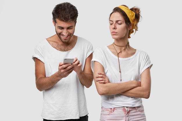 Бесплатное фото Ревнивая жена скрещивает руки, недовольно смотрит на мужа, который игнорирует ее, играет в онлайн-игры на мобильном телефоне, болтает с друзьями