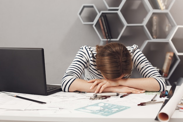 Free photo job occupation and overwork. close up of tired young good-looking engineer girl with dark hair in striped clothes lying on hands in office, suffering from headache after long work day.