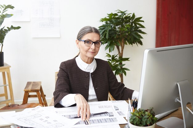 Job, occupation, profession and career concept. Attractive female architect in her fifties working on computer in office, calculating measurements and making drawings of construction project