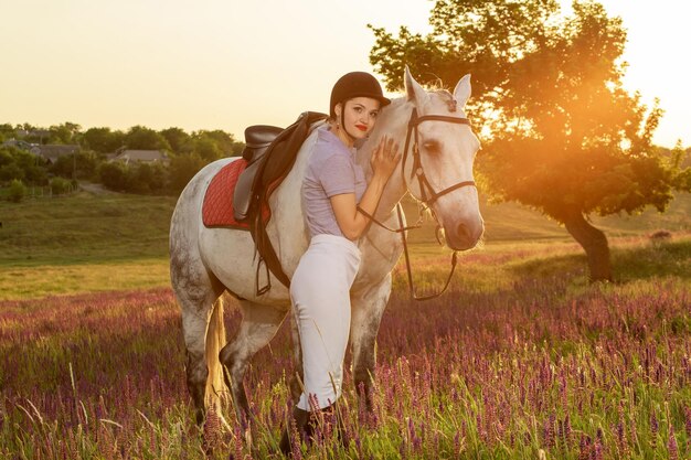 Маленькая девочка жокея гладит и обнимает белую лошадь в вечернем закате. Солнечная вспышка