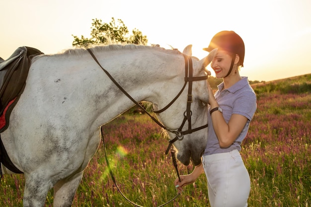 Jockey young girl petting and hugging white horse in evening sunset. Sun flare