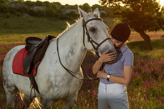 夕方の日没で白い馬をかわいがって抱き締めるジョッキーの若い女の子