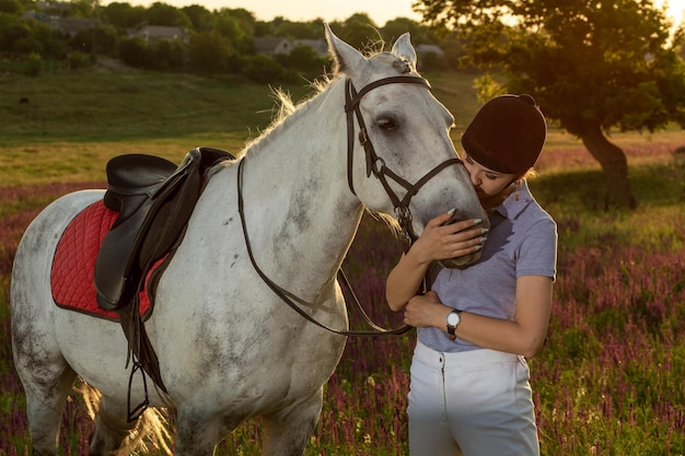 夕方の日没で白い馬をかわいがって抱き締めるジョッキーの若い女の子