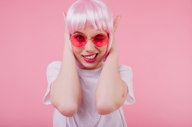 그녀의 귀를 덮고 선글라스에 Jocund 매력적인 여성 모델. 파스텔 벽에 고립 된 분홍색 머리를 가진 사랑스러운 소녀