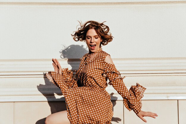 Jocund brown-haired woman dancing on the street. Portrait of emotional european girl in brown dress.