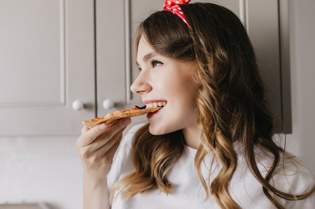 Jocund dark-haired woman enjoying fast food. Indoor close-up shot of elegant girl eating pizza.